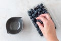 WomanÃ¢â¬â¢s hand picking blueberries from clamshell container on a white granite counter, small black bowl